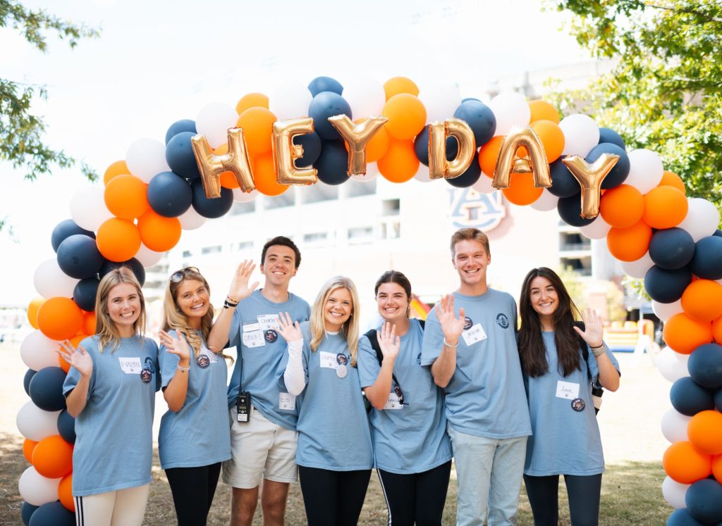 Hey Day Students smile and say "Hey" under an orange and blue balloon arch on the campus green
